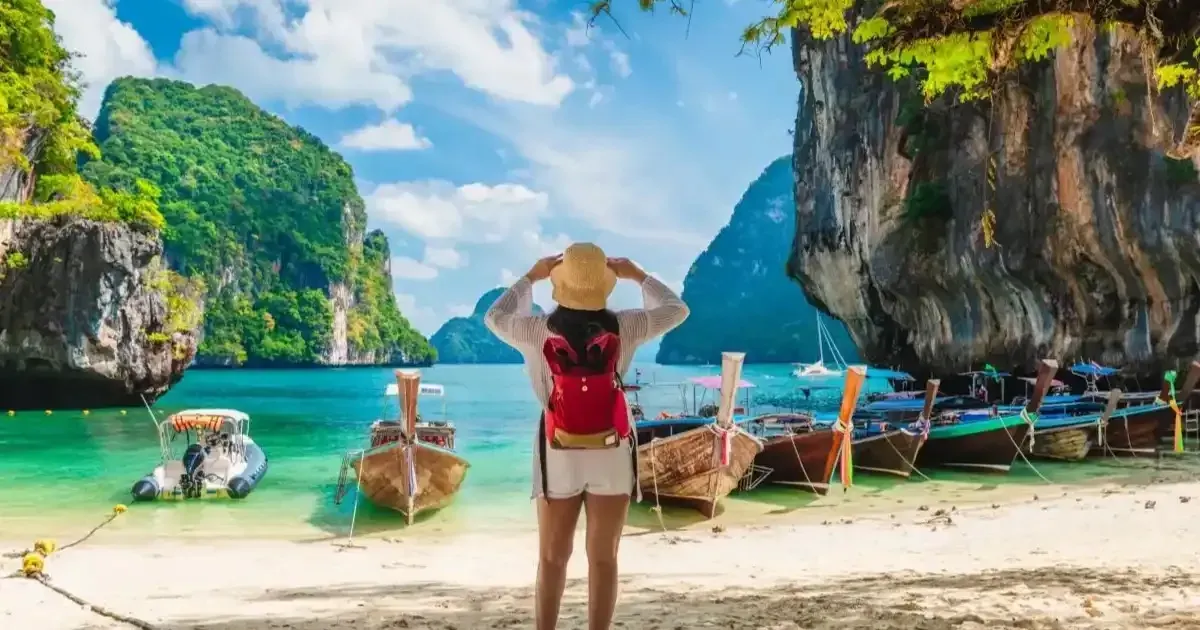 A traveller in a sun hat looking at longtail boats on a beach between limestone cliffs in southern Thailand